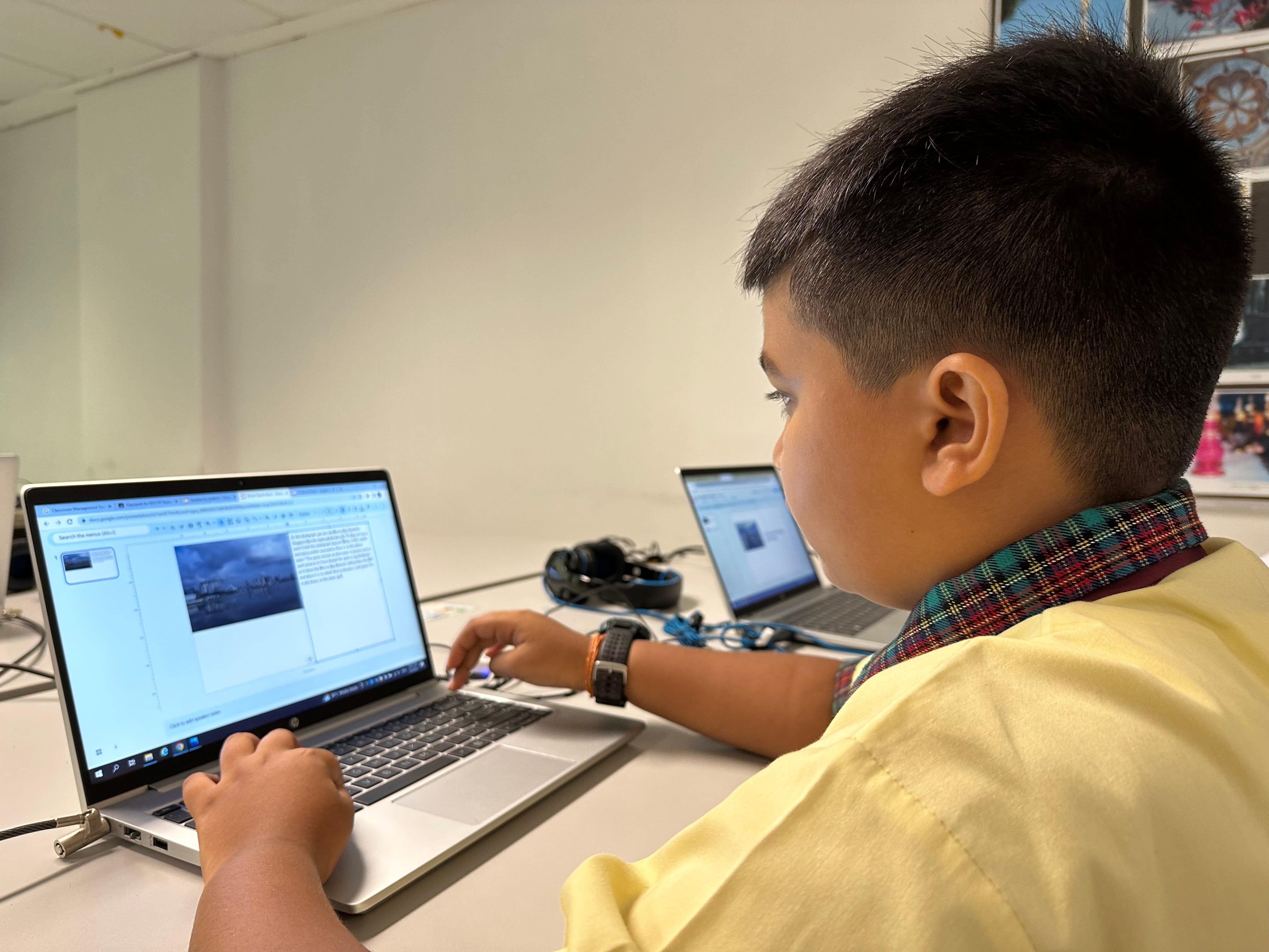 A young boy in a yellow shirt types on a laptop at a desk, with a second laptop and headphones nearby.